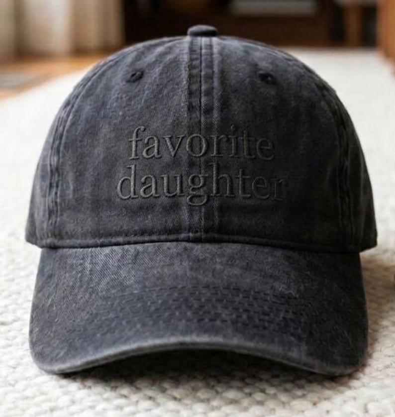 May include: A dark gray baseball cap with the embroidered text "favorite daughter" in a matching tone. The cap has a curved brim and a classic six-panel design, suitable for casual wear.
