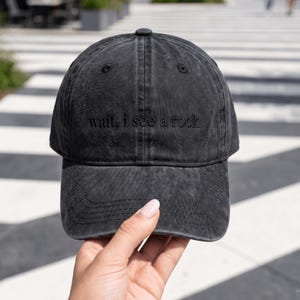 May include: A charcoal grey baseball cap with a distressed, washed-out finish. The front panel features the embroidered text "wait, i see a rock" in a matching dark thread. The cap has a structured crown, a curved brim, and stitched eyelets.