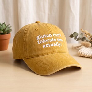 Peut inclure: Une casquette de baseball jaune moutarde avec une finition vieillie et un texte brodé en blanc sur le devant qui dit, gluten can't tolerate me, actually. La casquette présente une calotte structurée, une visière courbée et un bouton au sommet.