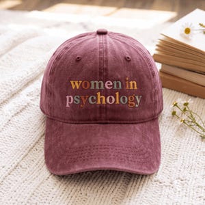 May include: A maroon baseball cap featuring the embroidered text "women in psychology" in multi-colored letters. The cap has a structured crown, a curved brim, and a button on top. The fabric has a slightly faded, washed-out appearance.