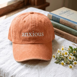 May include: A burnt orange baseball cap featuring the word anxious embroidered in white lowercase letters on the front panel. The cap has a structured crown, a curved brim with visible stitching, and a button at the top centre of the crown.