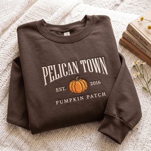 Puede incluir: Una sudadera de cuello redondo color marrón oscuro con texto bordado en blanco que dice PELICAN TOWN, EST. 2016 y PUMPKIN PATCH. Centrada entre el texto hay una calabaza naranja pixelada bordada con un tallo verde en el pecho.