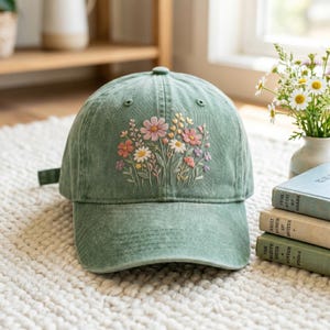 May include: A sage green baseball cap featuring an embroidered floral design on the front panel. The embroidery depicts a cluster of wildflowers in shades of pink, white, yellow, and purple with green stems and leaves on the cotton fabric.