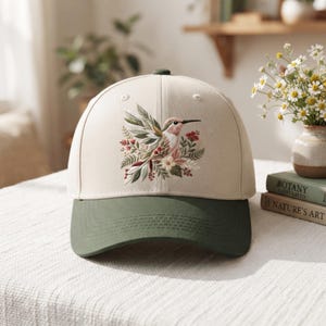 May include: A baseball cap with a cream-colored crown and a dark olive green brim. The front panel features an embroidered design of a hummingbird surrounded by green leaves, small red berries, and light-colored flowers in shades of pink and white.
