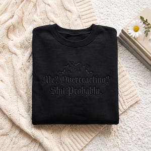 May include: A black crewneck sweatshirt featuring black embroidery of several small bats above the text "Me? Overreacting? Shit Probably." in a gothic-style font. The garment has a round neckline and long sleeves, with the design centered on the chest.