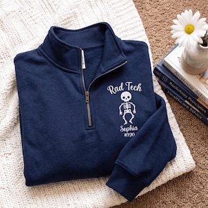 May include: A navy blue quarter-zip sweatshirt featuring a white graphic of a skeleton. The text reads Rad Tech, Sophia, and RT(R) in white lettering. The garment has a high collar with a metal zip closure and long sleeves with ribbed cuffs.