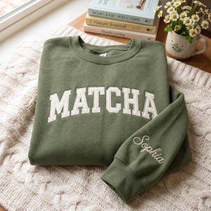 May include: An olive green crewneck sweatshirt featuring the word MATCHA in large, white, arched block letters across the chest. The sleeve is embroidered with the name Sophia in white cursive script, and the garment has a ribbed collar.