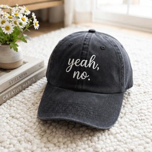 Puede incluir: Una gorra de béisbol gris carbón con el texto bordado en blanco "yeah, no." centrado en el panel frontal. La gorra tiene una construcción de seis paneles con visera curva y un botón en la parte superior, hecha de tela con aspecto desgastado.
