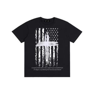 Puede incluir: Camiseta negra con un diseño de bandera estadounidense desgastada y una cruz blanca. El diseño de la bandera incorpora estrellas y rayas, con la cruz prominentemente en el centro. El texto "Congress shall make no law respecting an establishment of religion, or prohibiting the free exercise thereof" está impreso en la parte inferior.