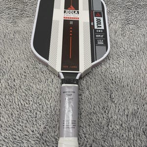 May include: A new JOOLA Pro Perseus table tennis paddle with a black and white design. The paddle has a white handle with a protective foil wrap that reads "REMOVE FOIL TAP HERE". The paddle is marked "USA" and "16 mm".