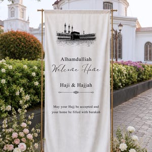 May include: A vertical off-white fabric banner suspended on a gold metal stand. The banner features a black ink illustration of the Kaaba, with text reading: Alhamdulliah, Welcome Home, Haji & Hajjah, May your Hajj be accepted and your home be filled with barakah.