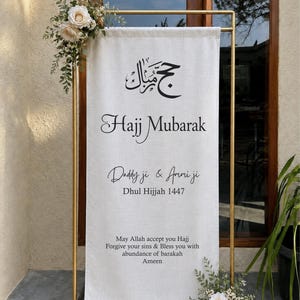 Puede incluir: Una pancarta vertical de tela color hueso colgada en un marco de metal dorado. La pancarta presenta texto negro que dice Hajj Mubarak, Daddy ji & Ammi ji, Dhul Hijjah 1447 y una oración. Rosas color crema y follaje verde están sujetos.