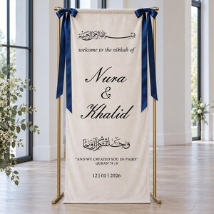 Pode incluir: Uma faixa de tecido cor de creme suspensa num suporte de metal dourado com fitas azul-escuras. A faixa apresenta texto a preto com "welcome to the nikkah of Nura & Khalid" e "AND WE CREATED YOU IN PAIRS" com a data 12|01|2026.