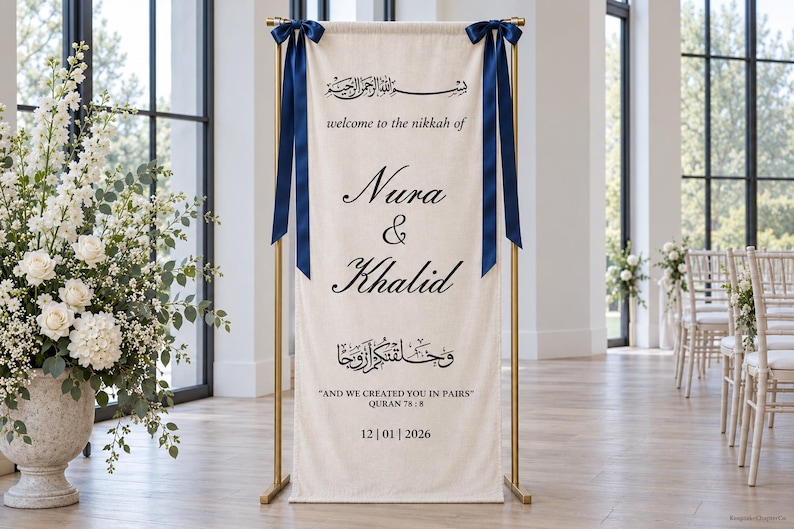 Pode incluir: Uma faixa de tecido cor de creme suspensa num suporte de metal dourado com fitas azul-escuras. A faixa apresenta texto a preto com "welcome to the nikkah of Nura & Khalid" e "AND WE CREATED YOU IN PAIRS" com a data 12|01|2026.