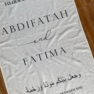 Pode incluir: Uma faixa de tecido retangular branca com texto impresso em preto que diz WELCOME TO OUR BEGINNING, ABDIFATAH and FATIMA, seguido de escrita &aacute;rabe e a tradu&ccedil;&atilde;o em ingl&ecirc;s AND HE PLACED BETWEEN YOU LOVE AND MERCY.