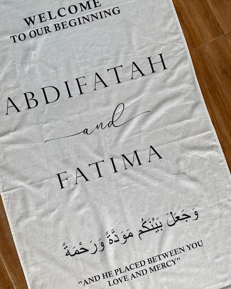 Pode incluir: Uma faixa de tecido retangular branca com texto impresso em preto que diz WELCOME TO OUR BEGINNING, ABDIFATAH and FATIMA, seguido de escrita &aacute;rabe e a tradu&ccedil;&atilde;o em ingl&ecirc;s AND HE PLACED BETWEEN YOU LOVE AND MERCY.