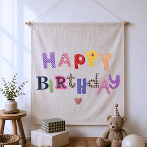 May include: A beige fabric wall hanging featuring the words HAPPY BIRTHDAY in colorful, patterned fabric letters. The letters are in various colors including purple, pink, red, yellow, orange, blue, and brown. A small red heart is below the text.