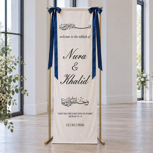 Pode incluir: Uma faixa alta e retangular de tecido em tom off-white suspensa por um suporte de metal dourado. A faixa apresenta la&ccedil;os de fita azul-escura nos cantos superiores e texto em preto com "welcome to the nikkah of Nura & Khalid" e "AND WE CREATED YOU IN PAIRS" com a data 12|01|2026.