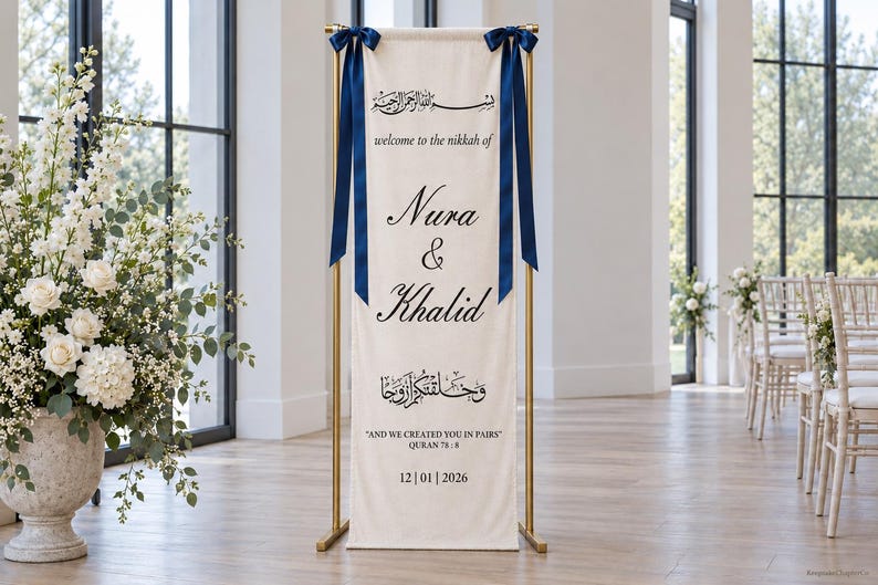 Pode incluir: Uma faixa alta e retangular de tecido em tom off-white suspensa por um suporte de metal dourado. A faixa apresenta la&ccedil;os de fita azul-escura nos cantos superiores e texto em preto com "welcome to the nikkah of Nura & Khalid" e "AND WE CREATED YOU IN PAIRS" com a data 12|01|2026.