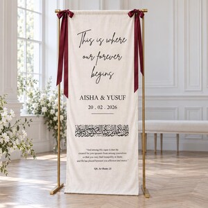 Pode incluir: Uma faixa vertical de tecido em tom off-white suspensa num suporte de metal dourado com fitas vermelho-escuras. A faixa apresenta o texto "This is where our forever begins," "AISHA & YUSUF," "20.02.2026," caligrafia &aacute;rabe e uma cita&ccedil;&atilde;o de QS. Ar-Rum: 21.