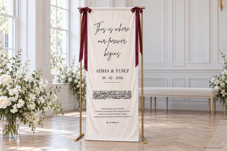 Pode incluir: Uma faixa vertical de tecido em tom off-white suspensa num suporte de metal dourado com fitas vermelho-escuras. A faixa apresenta o texto "This is where our forever begins," "AISHA & YUSUF," "20.02.2026," caligrafia &aacute;rabe e uma cita&ccedil;&atilde;o de QS. Ar-Rum: 21.