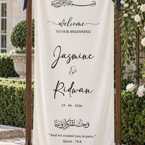 Pode incluir: Uma placa de boas-vindas de casamento em tecido creme pendurada numa moldura de madeira escura com fitas azul-marinho. O texto diz: Welcome to our beginning, Jasmine & Ridwan, 24.06.2026, e And we created you in pairs. Quran 78:8.