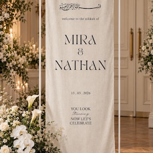 Pode incluir: Uma placa de casamento alta e retangular de tecido bege suspensa numa estrutura de metal branco. O texto diz: "welcome to the nikkah of MIRA & NATHAN 13.03.2026 YOU LOOK stunning NOW LET'S CELEBRATE" com caligrafia árabe no topo.