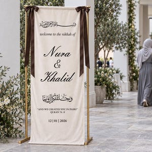 Pode incluir: Uma placa de boas-vindas de casamento em tecido cor de creme suspensa numa estrutura de metal dourado com fitas castanho-escuras. A placa apresenta texto a preto com Nura & Khalid, welcome to the nikkah of e And we created you in pairs.