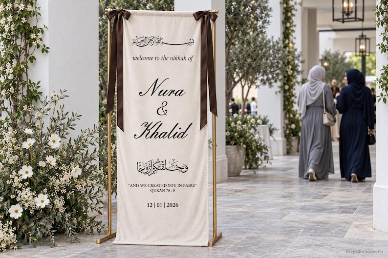Pode incluir: Uma placa de boas-vindas de casamento em tecido cor de creme suspensa numa estrutura de metal dourado com fitas castanho-escuras. A placa apresenta texto a preto com Nura & Khalid, welcome to the nikkah of e And we created you in pairs.