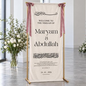 Pode incluir: Um estandarte de tecido cor de creme num suporte de metal dourado, com texto preto que diz Welcome to the Nikkah of Maryam & Abdullah, caligrafia &aacute;rabe, um vers&iacute;culo do Alcor&atilde;o e a data 24.01.2026, decorado com duas fitas rosa seco.