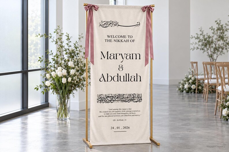 Pode incluir: Um estandarte de tecido cor de creme num suporte de metal dourado, com texto preto que diz Welcome to the Nikkah of Maryam & Abdullah, caligrafia &aacute;rabe, um vers&iacute;culo do Alcor&atilde;o e a data 24.01.2026, decorado com duas fitas rosa seco.