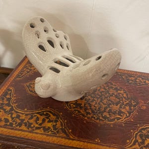 May include: A cream-colored ceramic butterfly figurine with a crackle glaze finish. The butterfly has openwork wings and a rounded body. The figurine is displayed on a dark brown, patterned surface.