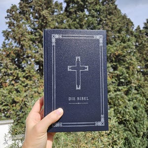 Könnte beinhalten: Ein dunkelblaues Buch mit dem Titel "DIE BIBEL" und einem silbernen Kreuzdesign. Das Buch hat einen dekorativen Rahmen. Der Hintergrund ist eine verschwommene Ansicht von grünen Bäumen.