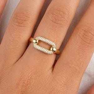 May include: A gold ring featuring a rectangular link design, adorned with small, clear gemstones. The ring is on a finger, set against a white background. The ring's design is a modern take on a classic style.