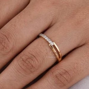 May include: A gold ring with a double band design. The top band is set with a row of small, clear gemstones. The ring is on a finger, highlighting its delicate and elegant style. The ring is a piece of jewellery.