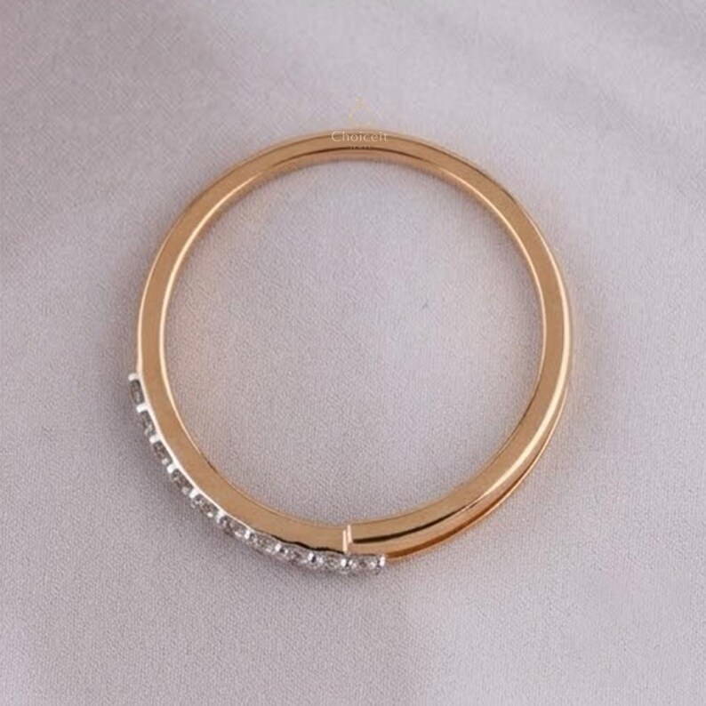 May include: A gold-coloured ring featuring a row of small, clear gemstones. The ring is circular and rests on a light-coloured, textured surface. The ring's design is simple and elegant, suitable for everyday wear.