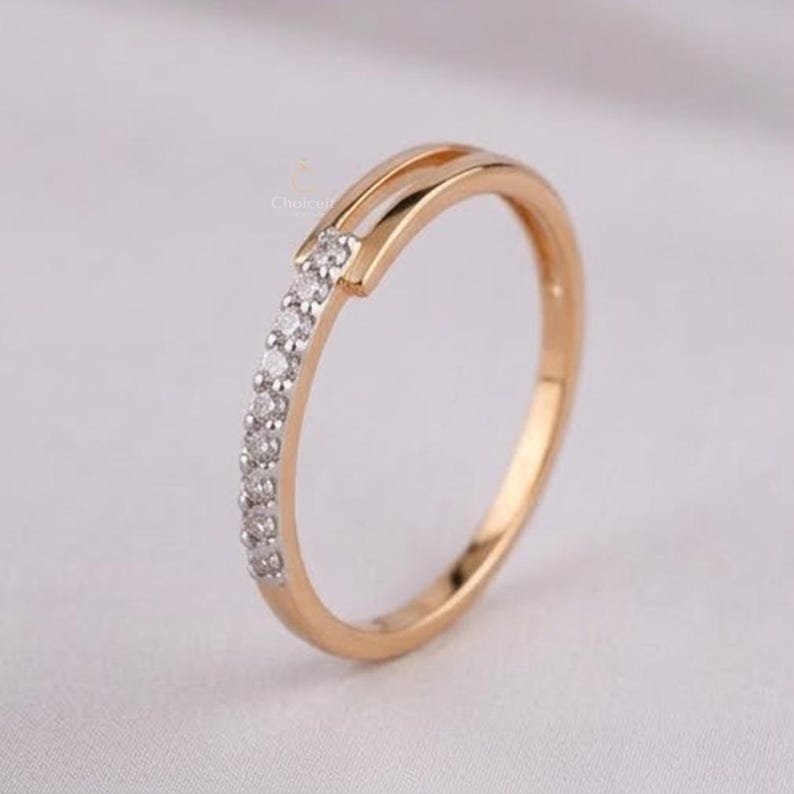 May include: A gold ring featuring a line of small, clear gemstones along one side. The ring has a split design at the top, adding a distinctive touch. The ring is likely made of gold or a gold-toned metal.