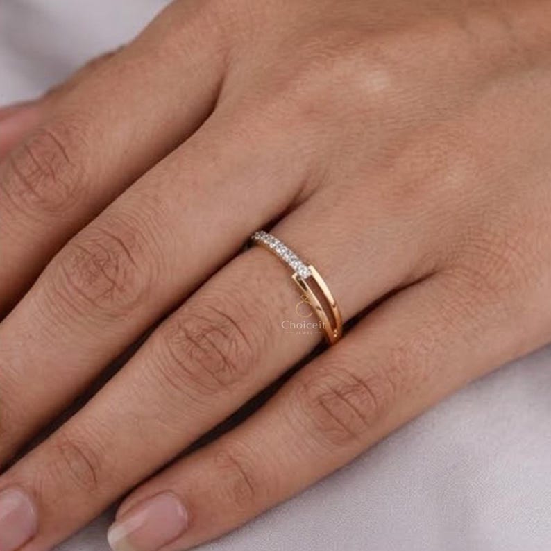 May include: A gold ring with a double band design. The top band is set with a row of small, clear gemstones. The ring is on a finger, highlighting its delicate and elegant style. The ring is a piece of jewellery.
