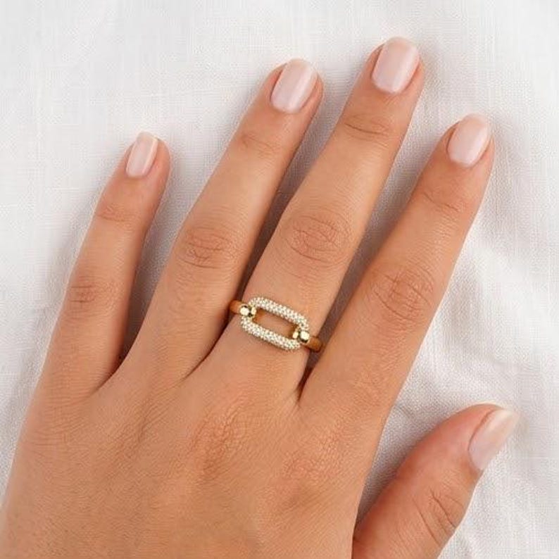 May include: A gold ring featuring a rectangular link design, adorned with small, clear gemstones. The ring is on a finger, set against a white background. The ring's design is a modern take on a classic style.
