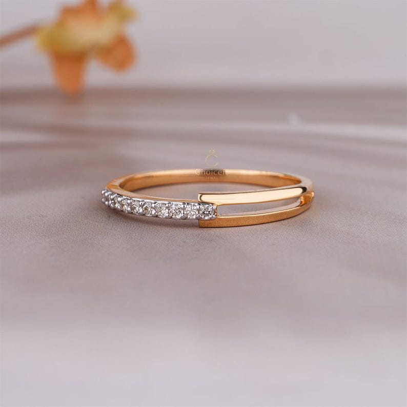 May include: A gold ring featuring a line of small, clear gemstones along one edge. The ring has a distinctive design with a straight, parallel bar on the other side. The ring is presented on a neutral, light-coloured surface.