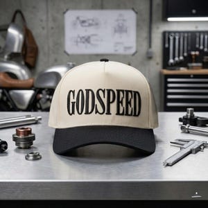 May include: A beige and black baseball cap with the word "GODSPEED" embroidered in black across the front. The cap has a black brim and a black button on top. The cap is sitting on a metal surface.
