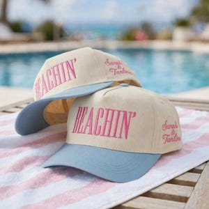 May include: Two cream-colored baseball caps with light blue brims. The front of each cap features the word BEACHIN' embroidered in pink. On the side of each cap, the text Sarah & Tanlines is embroidered in pink script lettering.