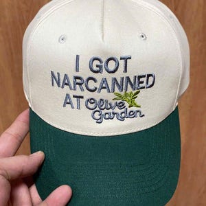 May include: A cream-colored baseball cap with a dark green brim and a dark green button on top. The front features embroidered text that reads I GOT NARCANNED AT Olive Garden, with a small green olive branch graphic next to the word Olive.