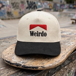 May include: A baseball cap with an off-white crown and a black brim. The front features an embroidered red chevron shape above the black text "Weirdo" in a serif font. The cap has a small black button at the top center of the crown.