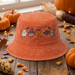 May include: An orange bucket hat with a washed-out texture featuring a row of embroidered Halloween icons including a white ghost, a purple witch hat, a smiling jack-o'-lantern, a black bat, and a gray tombstone with the letters RIP.