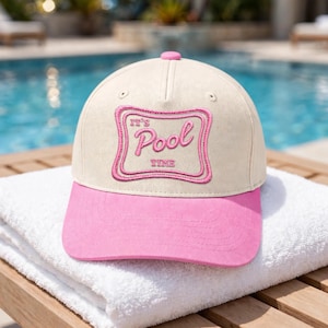 May include: A baseball cap with a cream-colored crown and a bright pink brim. The front features a rectangular embroidered patch with a pink border and the text "IT'S Pool TIME" in pink script and block lettering, centered on the front panel.