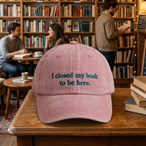 May include: Dusty rose baseball cap with the embroidered text "I closed my book to be here." The hat is made of a soft, textured material and features a curved brim and adjustable strap. The hat is displayed on a wooden table in a library setting.