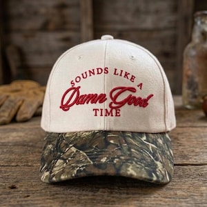 May include: A cream-colored baseball cap with a camouflage print brim. The front of the cap features red embroidered text that reads, "SOUNDS LIKE A Damn Good TIME" in a mix of block and cursive lettering, centered on the crown of the hat.