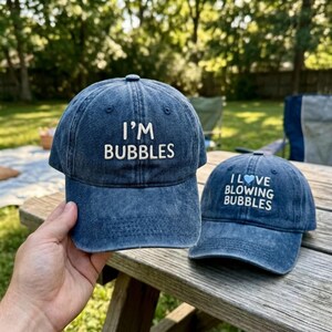 May include: Two blue denim baseball caps with white embroidered text. The larger cap features the text I'M BUBBLES. The smaller cap features the text I LOVE BLOWING BUBBLES with a light blue heart icon replacing the word LOVE.