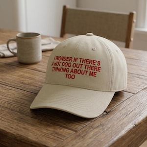 May include: Tan baseball cap with the text "I WONDER IF THERE'S A HOT DOG OUT THERE THINKING ABOUT ME TOO" in red. The cap is on a wooden table, with a mug and chair in the background.